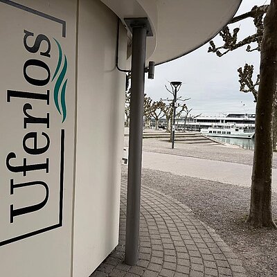 Der Uferlos-Kiosk mit Logo, dahinter der Konstanzer Hafen