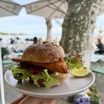 Fischburger mit Salat, dahinter Sonnenschirme und der See